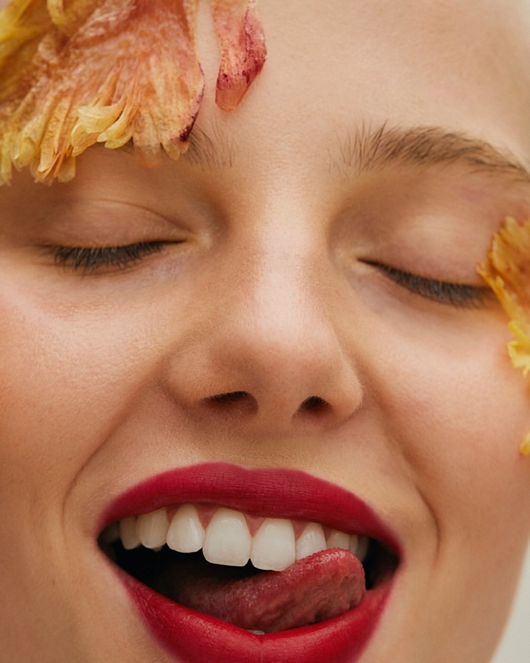 Close-up of a young woman with closed eyes and red lipstick, tongue sticking out, and flower petals around her face.