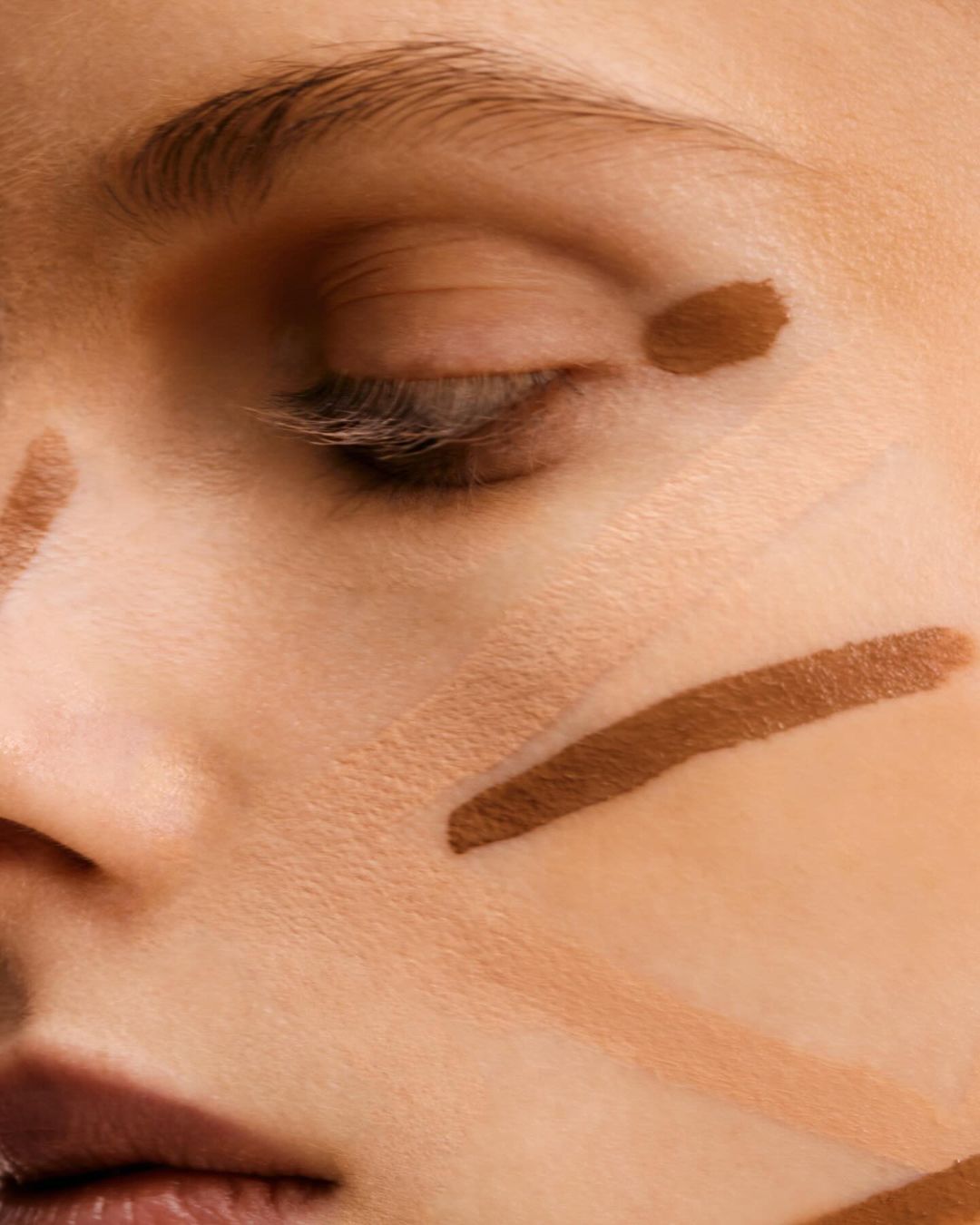 Close-up of a woman's face with makeup contour lines on her cheek, nose, and under the eye, shot in a brightly lit indoor setting.