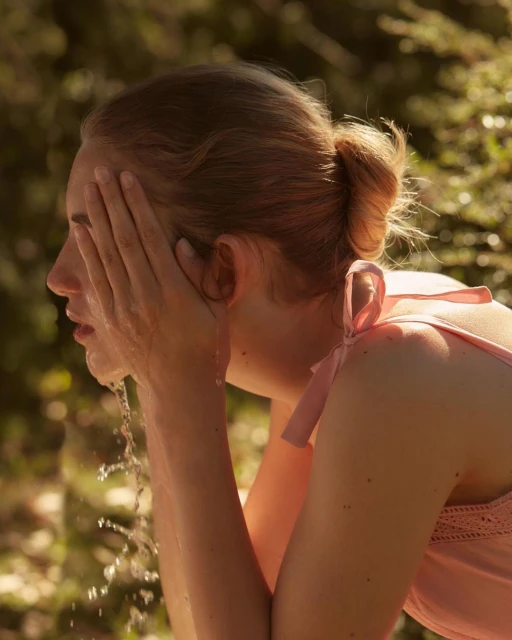 Woman washing her face outdoors in warm light, water splashing, hair in a bun, sleeveless top, green bokeh background.