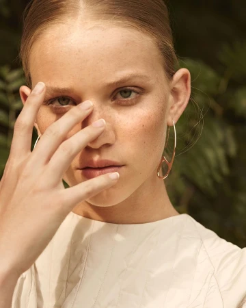 Close-up portrait of a woman with freckles holding her hand near her face wearing a gold earring