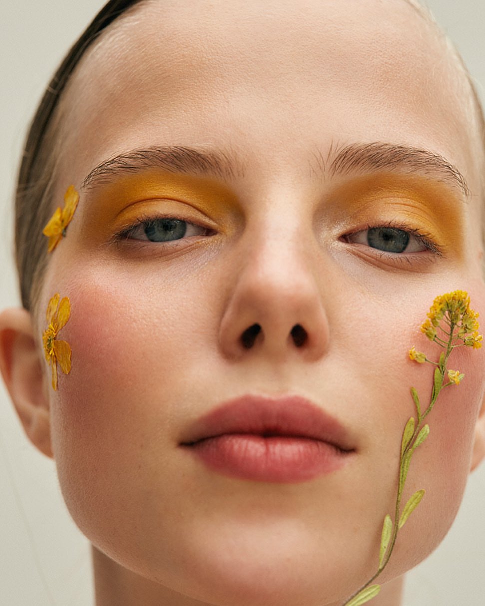 Close-up studio shot of a young adult woman with yellow eyeshadow, flowers on her face, and fair skin.