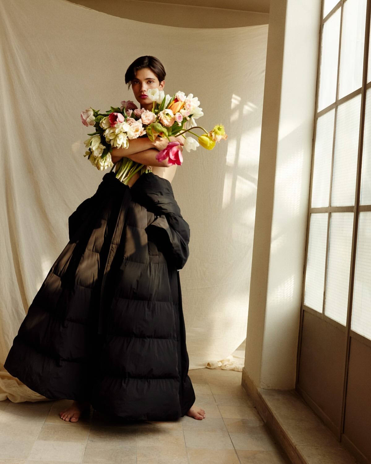 Woman poses with vibrant tulips and a voluminous black puffer coat indoors by a window, bathed in warm natural lighting.