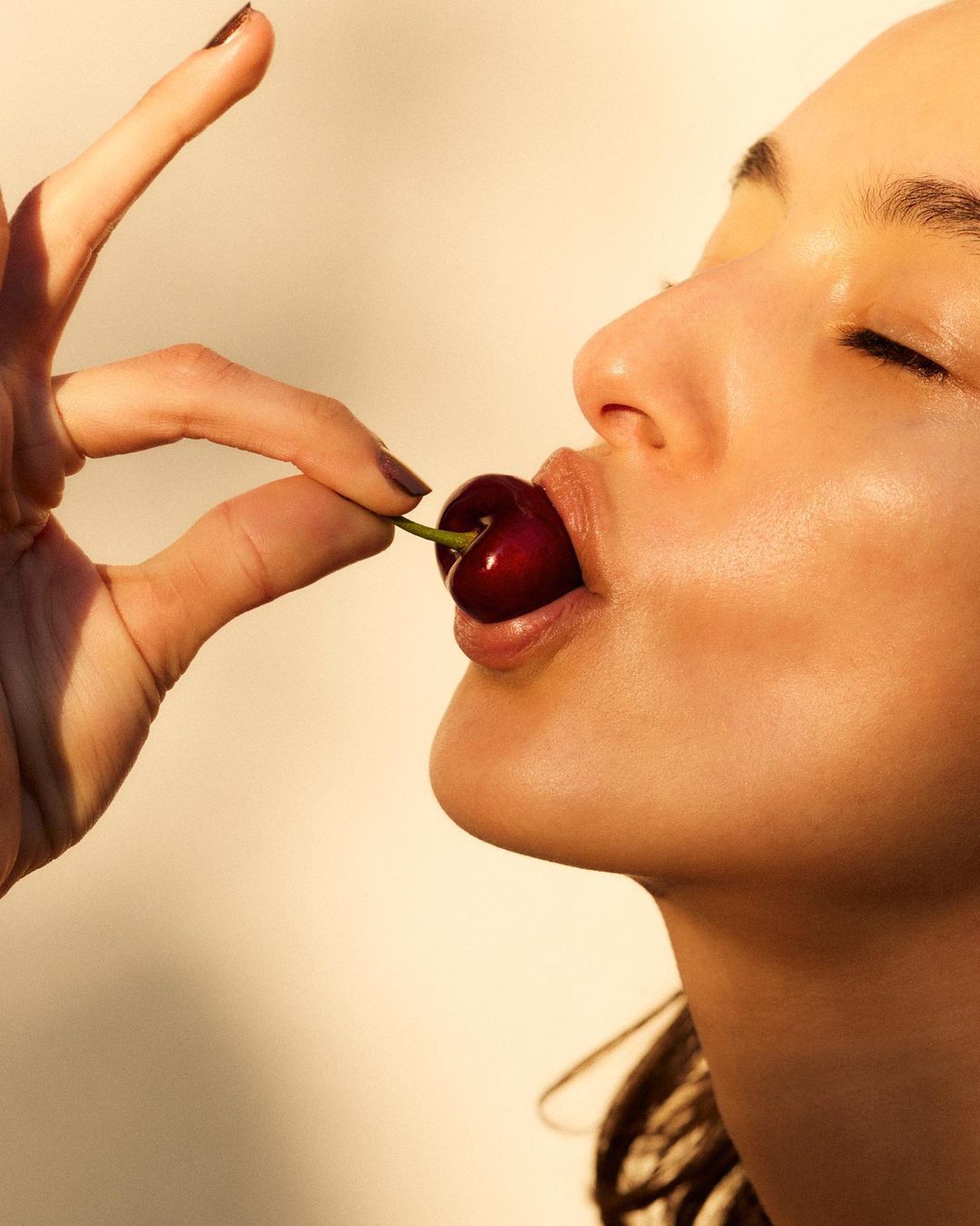 Close-up studio shot of an adult female model with a single cherry on her tongue, eyes closed, in a beauty and fashion context.