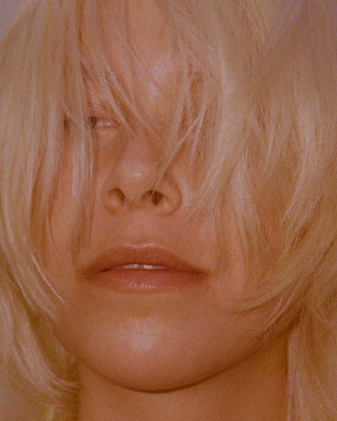 Close-up portrait of a blonde woman with hair partially covering her face, soft lighting, warm tones, and a blurred background.