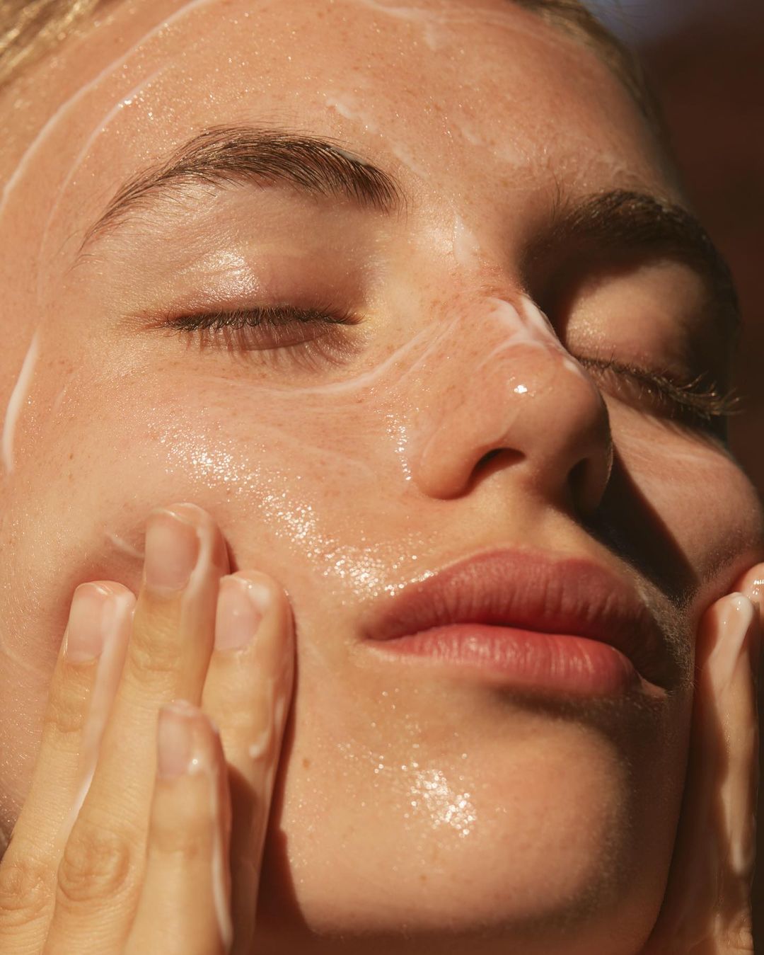 Close-up of an adult female with eyes closed, applying a foamy cleanser to her face. Natural lighting enhances the skin's texture.