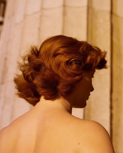 Profile view of an auburn-haired woman with bare shoulders against a background of blurred architectural columns, focusing on hair texture and colour.