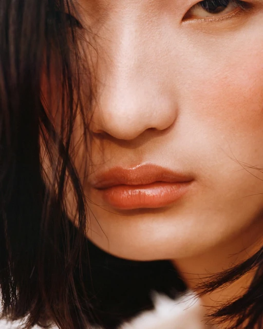 Close-up portrait of a young adult female showcasing beauty makeup with glossy lips and dark hair in soft, natural lighting.