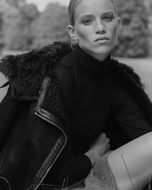 Monochrome portrait of a fair-skinned woman in a dark turtleneck and sheepskin jacket, seated in a grassy field, gazing at the camera.