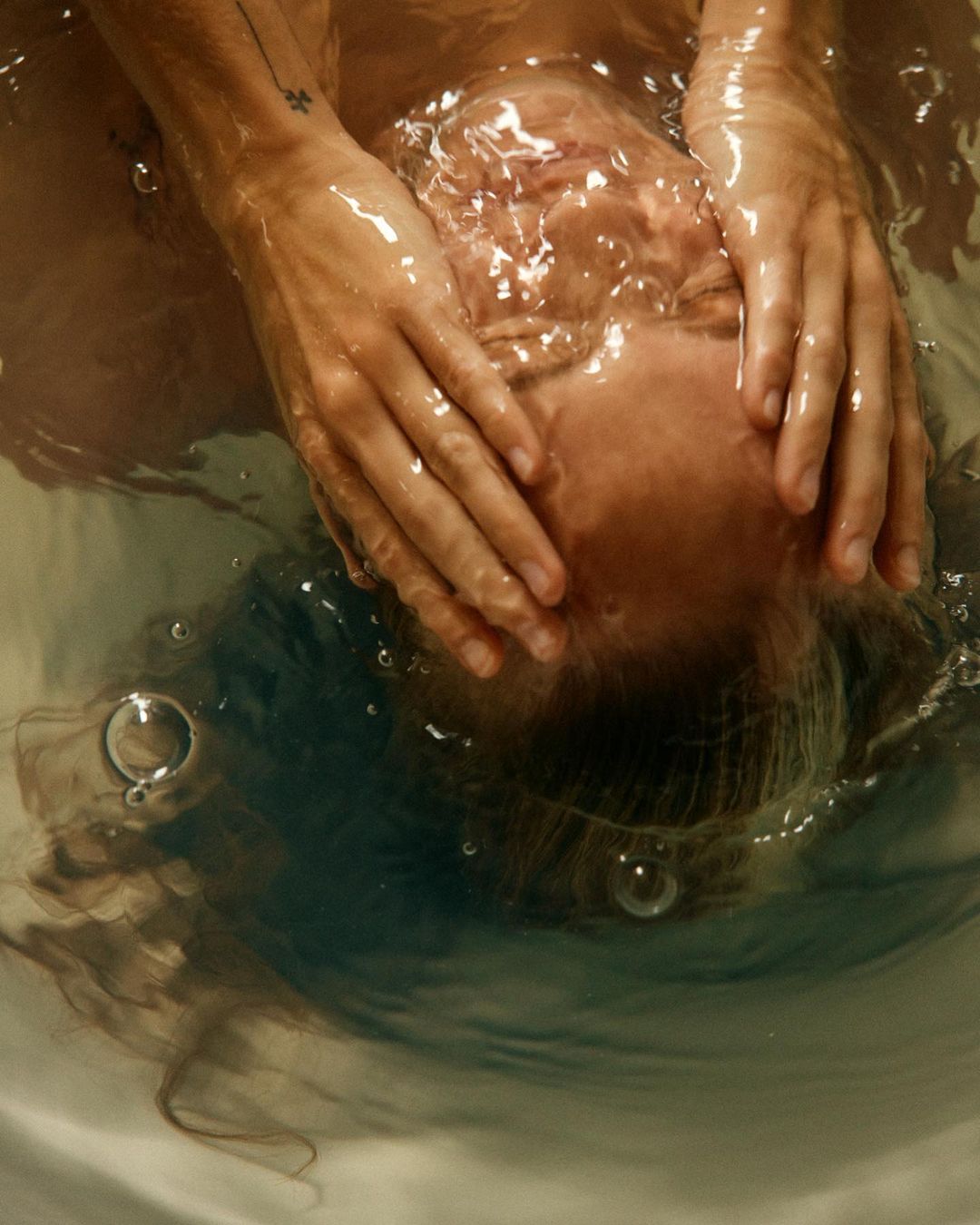 Overhead shot of person's face under water with hands over their head in bathtub. Visible bubbles and hair floating in the water.
