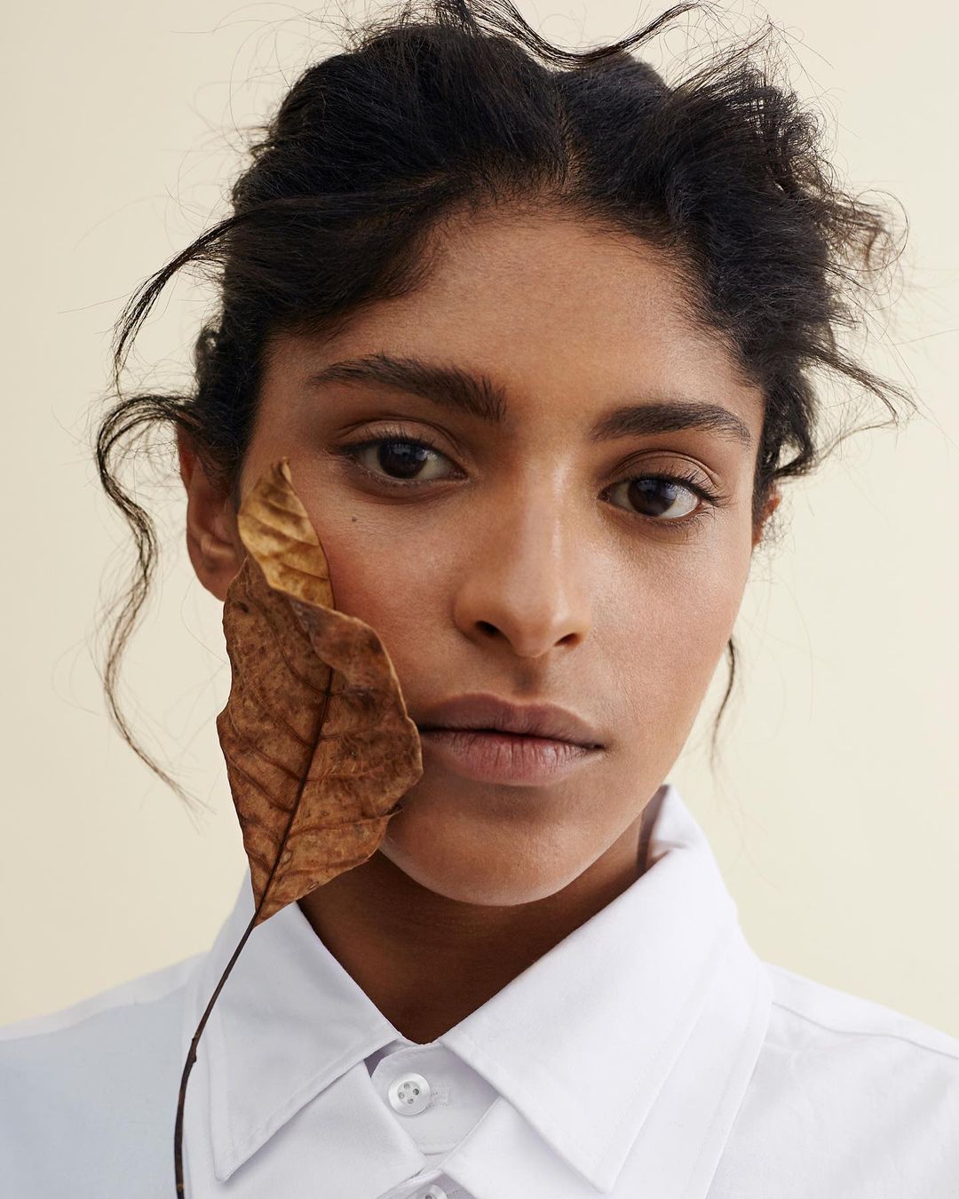 Close-up portrait of a woman with dark hair, wearing a white shirt, with a large dried leaf partially covering her face.
