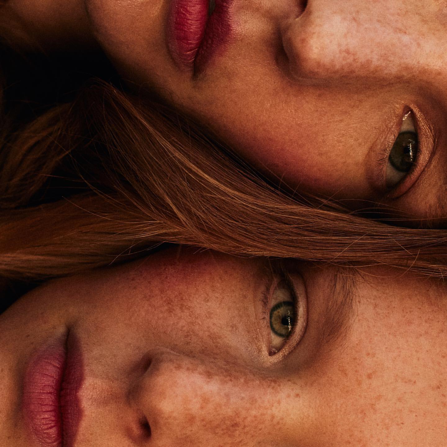 Close-up portrait of two fair-skinned adult females with freckles, red lipstick, and reddish-brown hair, faces positioned head-to-head.