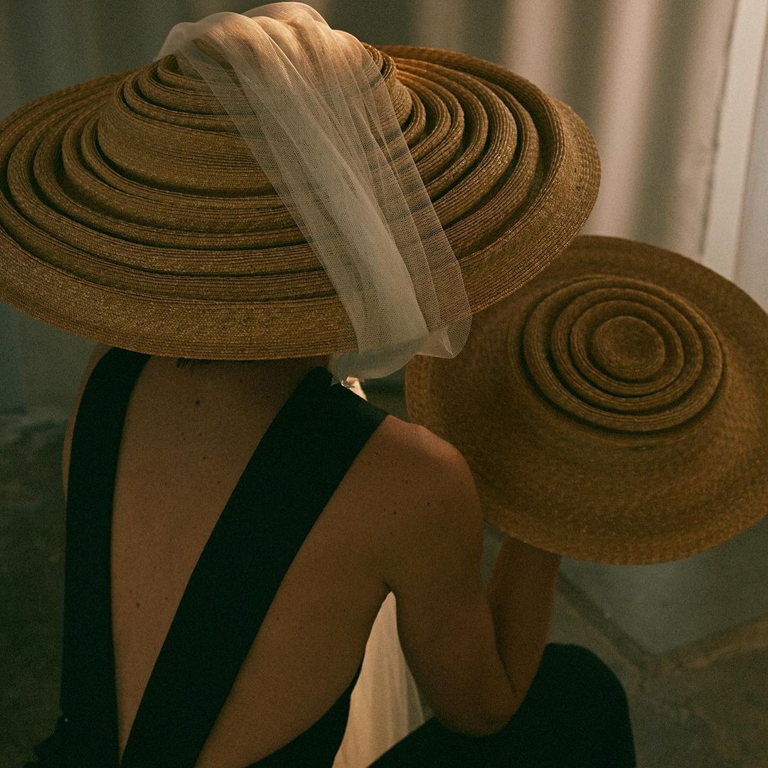 Two women in black dresses and large straw hats, one with a white veil, in a softly lit indoor setting, showcase vintage-inspired fashion.