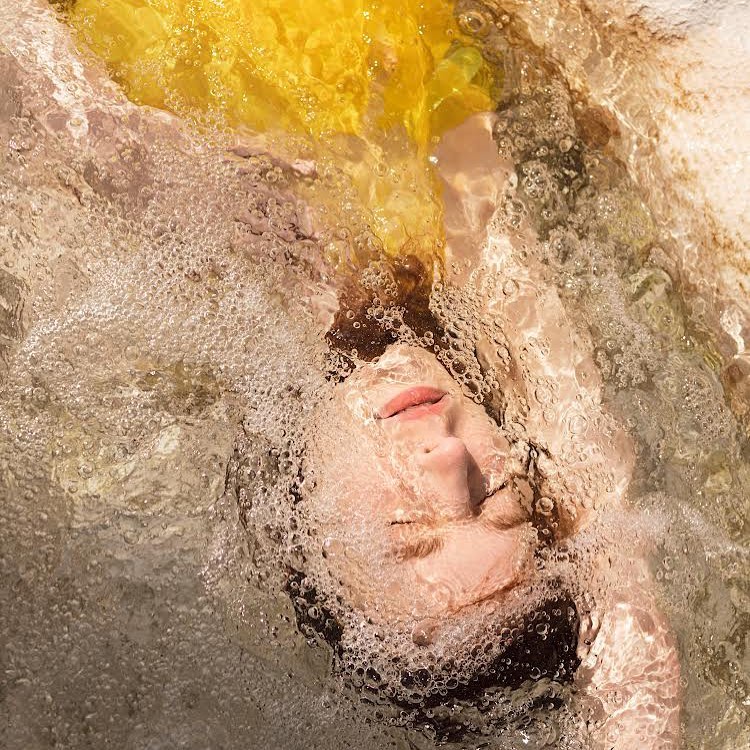 Portrait of woman underwater with flowing yellow fabric, creating a dreamlike effect with bubbles and diffused light in an indoor pool.