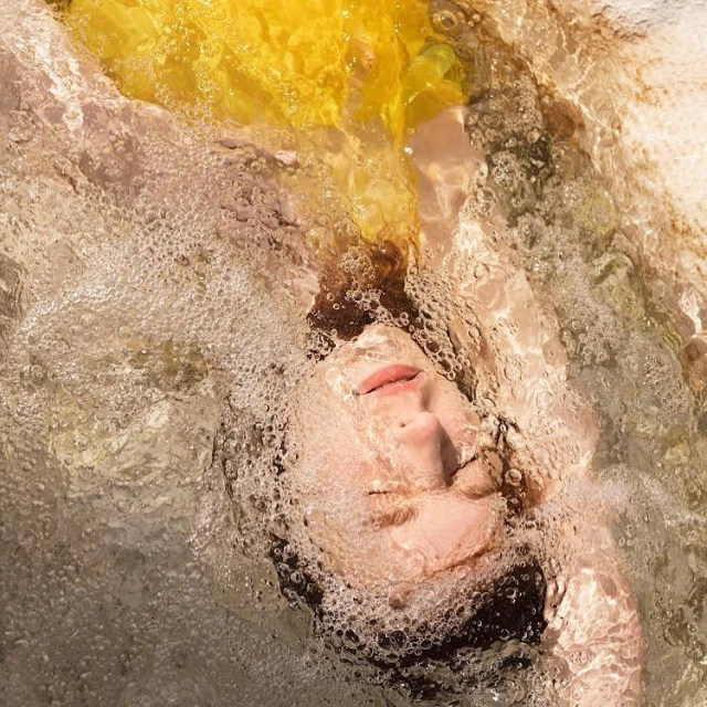Portrait of woman underwater with flowing yellow fabric, creating a dreamlike effect with bubbles and diffused light in an indoor pool.
