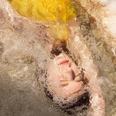 Portrait of woman underwater with flowing yellow fabric, creating a dreamlike effect with bubbles and diffused light in an indoor pool.