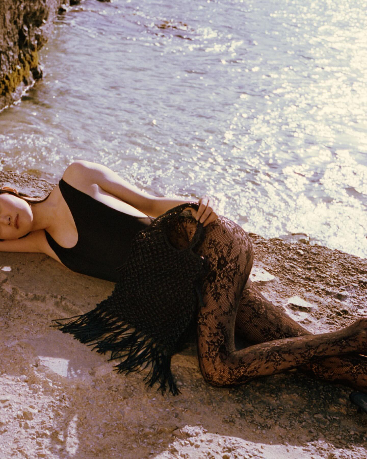 A woman in a black swimsuit lies on the sand near the ocean, partially covered by a dark fringed shawl and floral lace leggings.