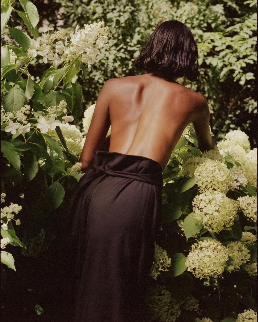Woman with bare back stands amongst white hydrangea blossoms, facing away from the camera on an overcast day in a lush garden.