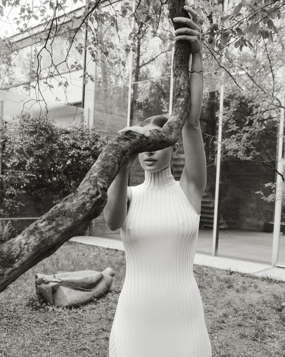 Monochrome portrait of a woman in a knit dress, posing with her hands on a tree branch, set against a modern building and leafy trees.