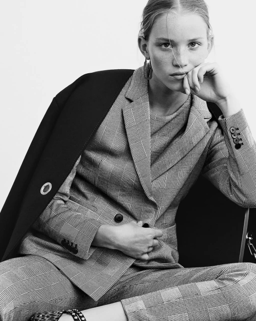 Monochrome fashion portrait of a female model wearing a plaid suit with a black coat draped over her shoulder, against a light background.