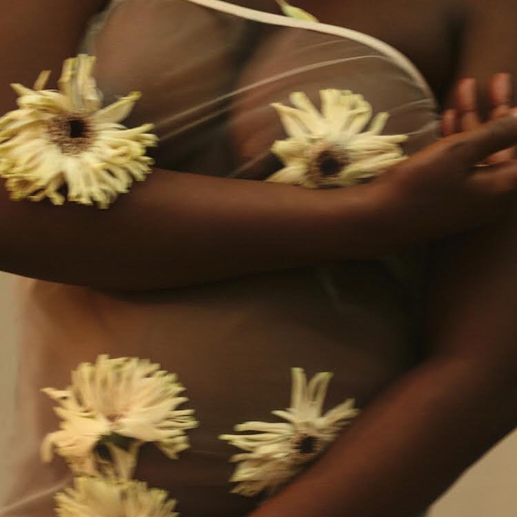 Upper torso shot of an adult woman with brown skin and sheer white top, posed with white and yellow flowers on arms and chest.