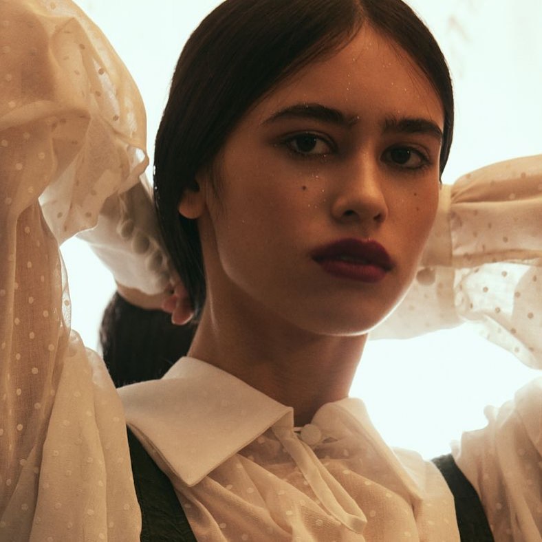 Close-up portrait of a young woman with dark hair and burgundy lipstick wearing a sheer white blouse and a black vest.