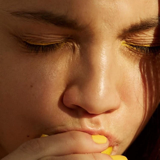 Close up of a face with fair skin and yellow eyeliner with an adult female eating a yellow fruit, bathed in warm, directional light.
