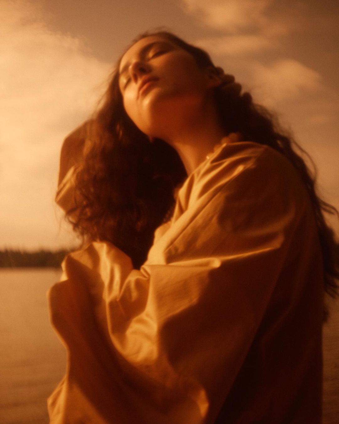 Portrait of a woman with long brown hair, wearing a beige shirt, posed in front of a lake and cloudy sky during golden hour.
