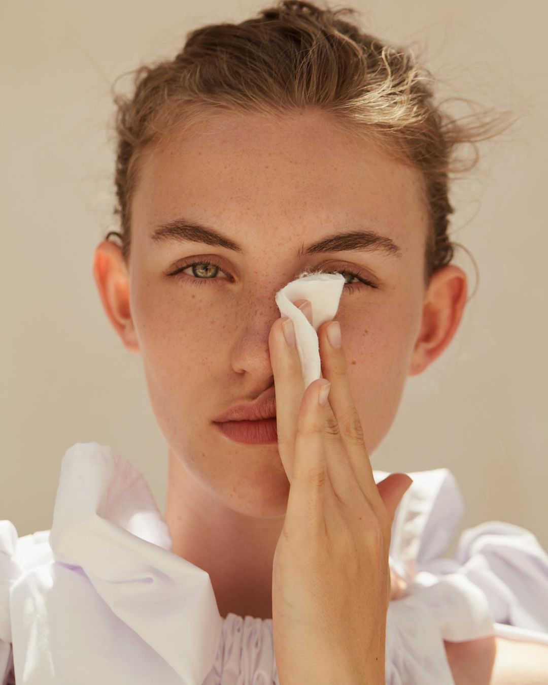 Close-up portrait of a young woman with fair skin and green eyes, holding a white cotton pad to her face, wearing a white ruffled shirt.
