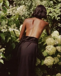 Topless woman with dark bob hair wearing dark trousers in a lush garden with white flowering hydrangeas.
