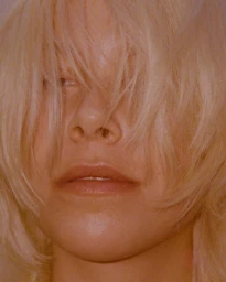 Close-up portrait of a person with long blonde hair partially covering their face and warm reddish lighting.