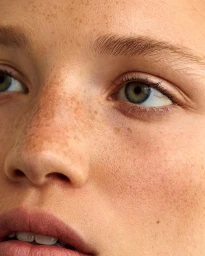 Close-up portrait of a woman face focusing on green eyes and detailed skin texture with freckles across her nose and cheeks.