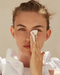 Close-up portrait of a woman holding a white tissue near her eye, wearing a ruffled white garment against a neutral backdrop.