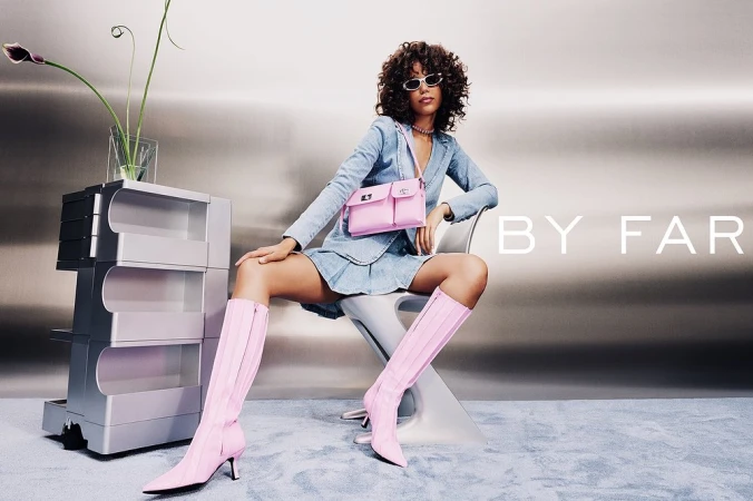 Woman in denim suit wearing pink boots and carrying a pink handbag, seated on a modern chair against a metallic gradient backdrop.