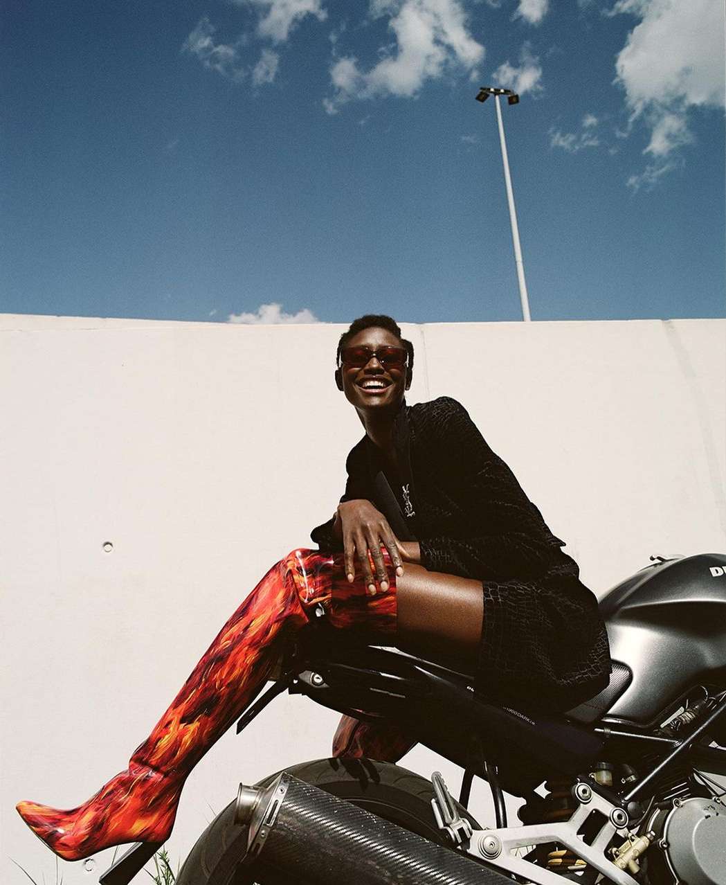 A dark-skinned woman in sunglasses and thigh-high flame print boots sits on a motorcycle against a white wall and blue sky.