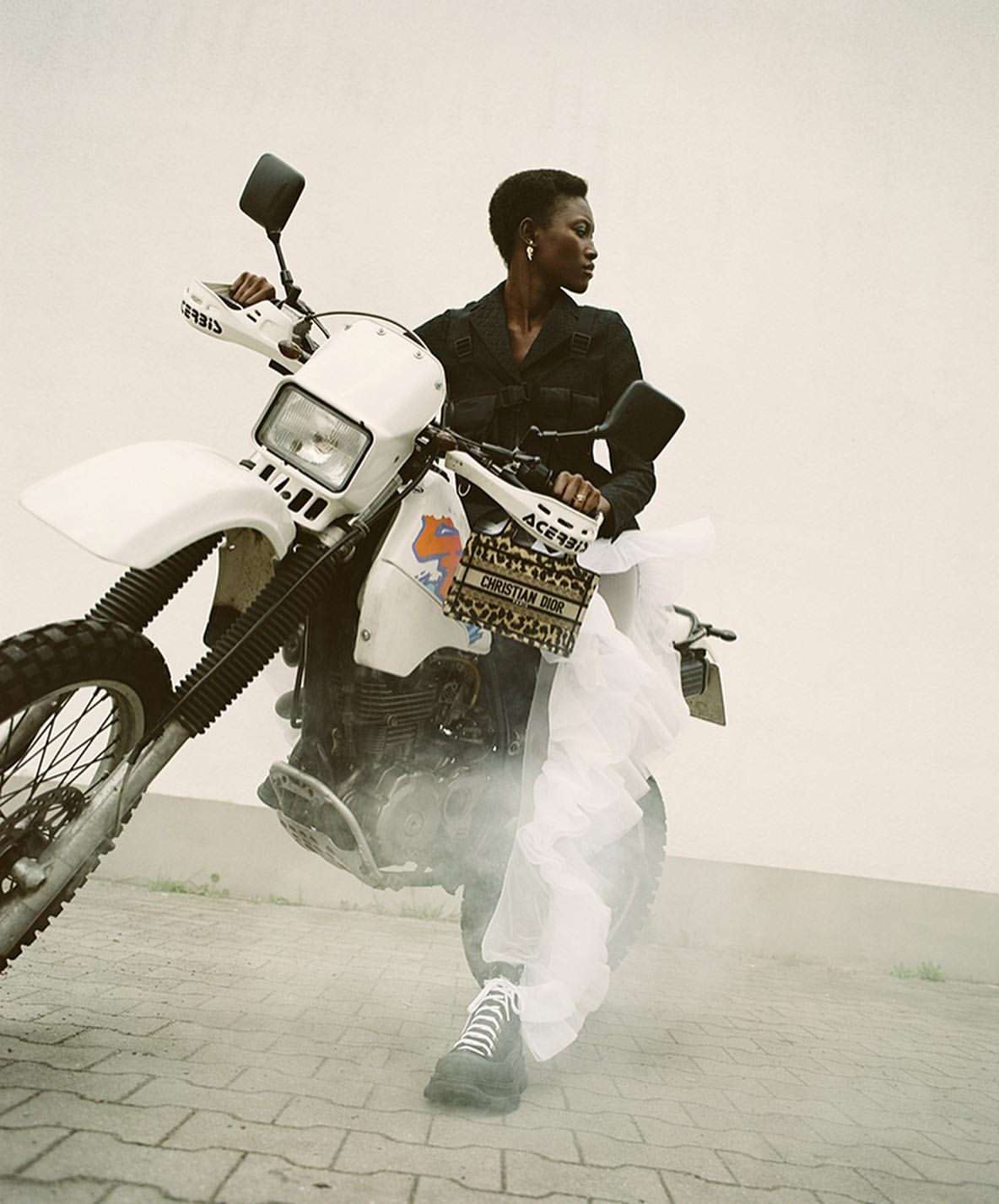 An adult Black woman in black clothing poses with a white dirt bike on a paved surface, accented by a designer handbag and tulle.