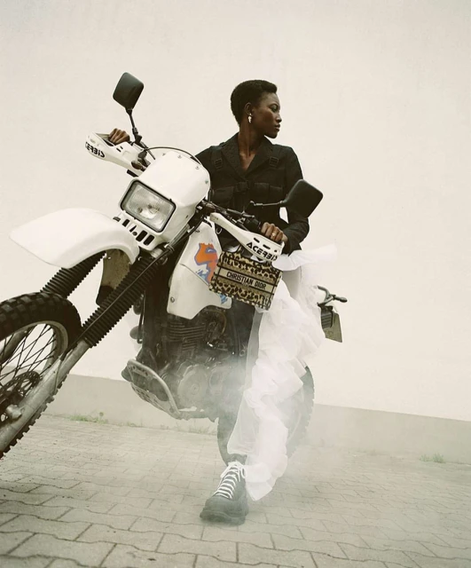 An adult Black woman in black clothing poses with a white dirt bike on a paved surface, accented by a designer handbag and tulle.