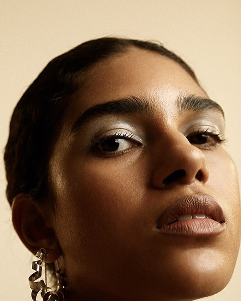 Portrait of a young adult female model with silver eyeshadow and hoop earrings, captured in a low-angle shot with a neutral background.