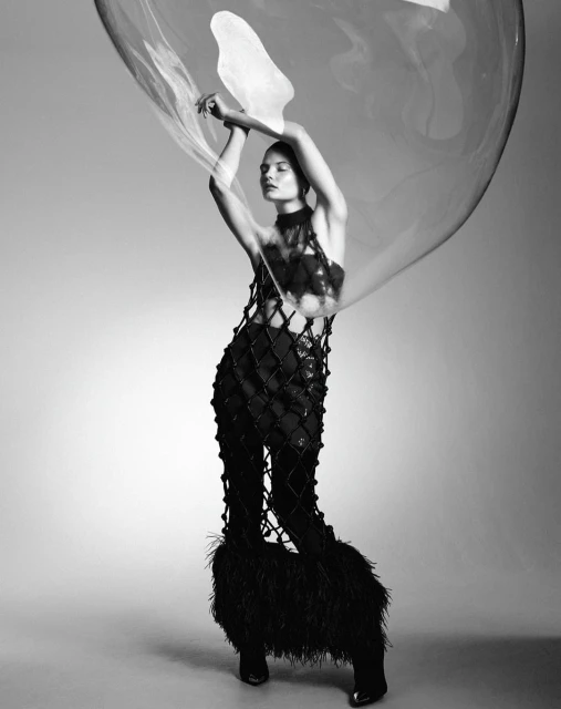 Monochrome fashion shot: Woman in a netted outfit poses with a large transparent sphere, creating an abstract portrait in a studio.