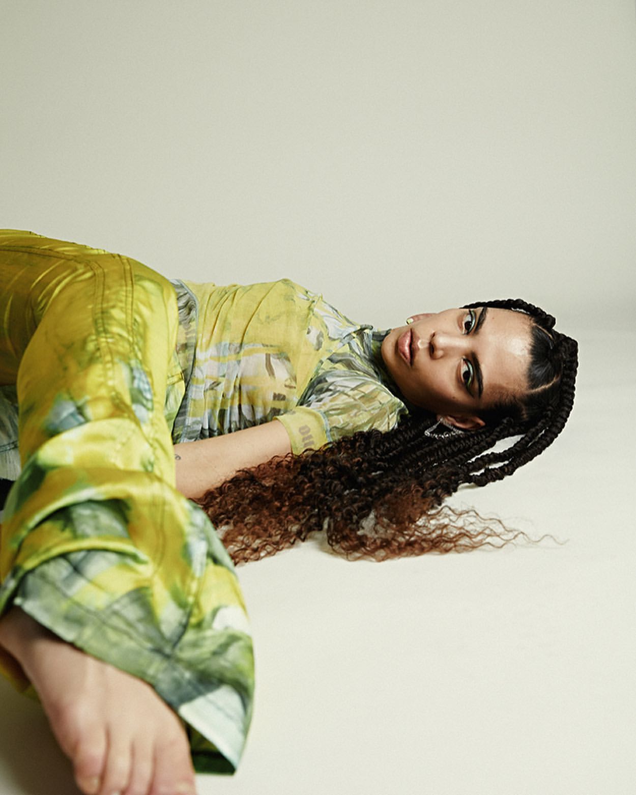 A young adult woman with braided dark hair poses in a green patterned outfit on a white surface, lying down and looking at the camera.