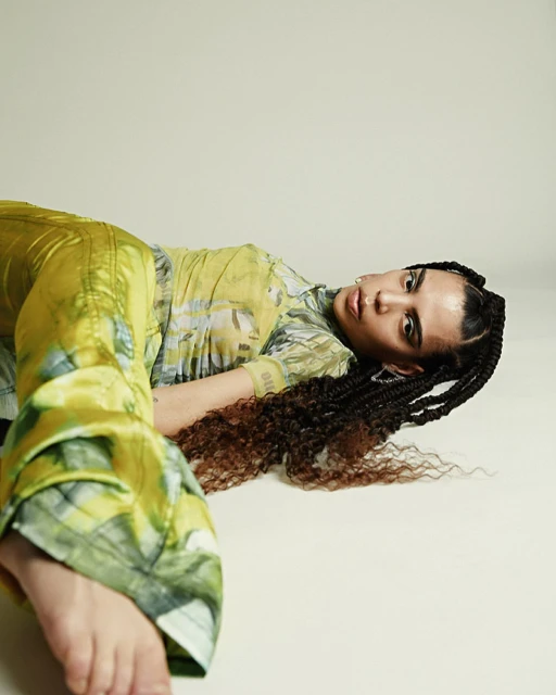 A young adult woman with braided dark hair poses in a green patterned outfit on a white surface, lying down and looking at the camera.