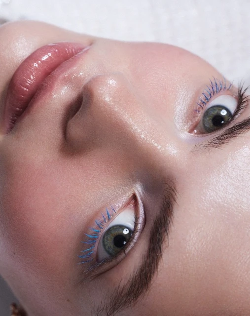 Close-up of a woman's face with light skin, green eyes, blue mascara, and glossy lips against a soft white textured background. 