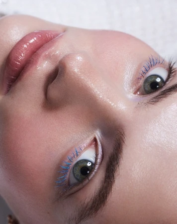 Close-up of a woman's face with light skin, green eyes, blue mascara, and glossy lips against a soft white textured background. 
