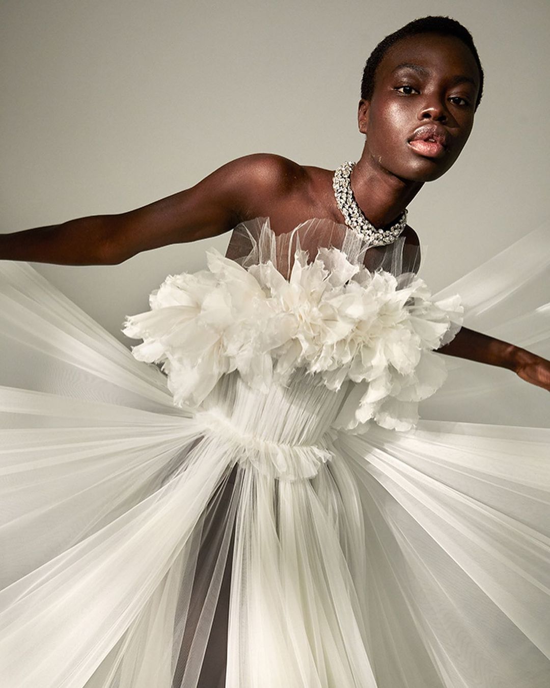 Elegant portrait of a woman in a white tulle gown and diamond necklace, striking a pose against a neutral backdrop.