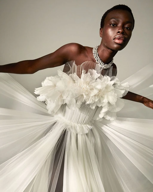 Elegant portrait of a woman in a white tulle gown and diamond necklace, striking a pose against a neutral backdrop.