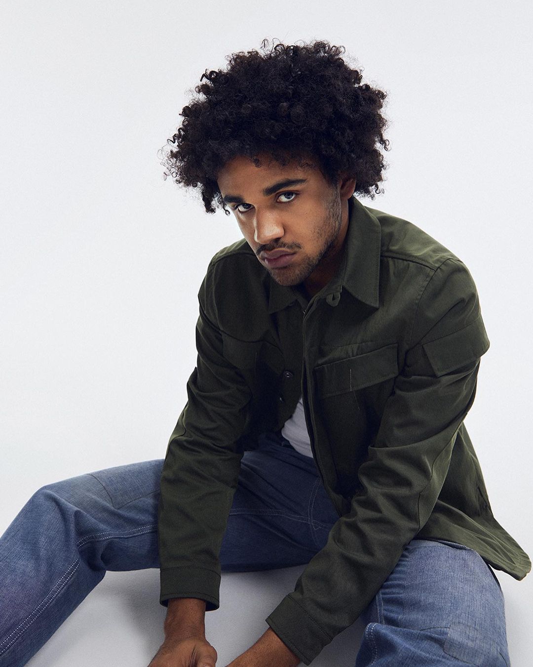 Portrait of a young adult male with an olive-green button-up shirt and blue jeans, seated against a bright white backdrop in a studio setting.