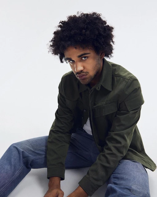 Portrait of a young adult male with an olive-green button-up shirt and blue jeans, seated against a bright white backdrop in a studio setting.