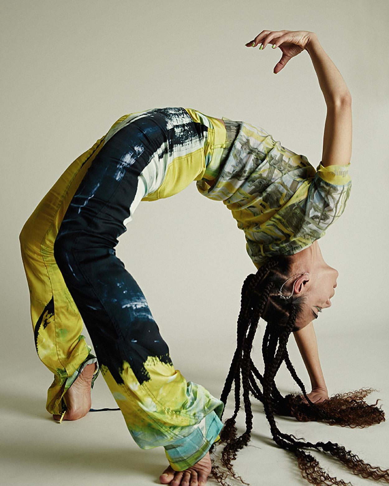 Woman in tie-dye pants and top strikes a backbend pose against a neutral backdrop, showcasing an athletic stretch.