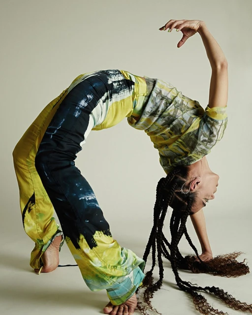 Woman in tie-dye pants and top strikes a backbend pose against a neutral backdrop, showcasing an athletic stretch.