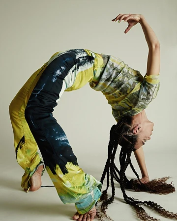 Woman in tie-dye pants and top strikes a backbend pose against a neutral backdrop, showcasing an athletic stretch.