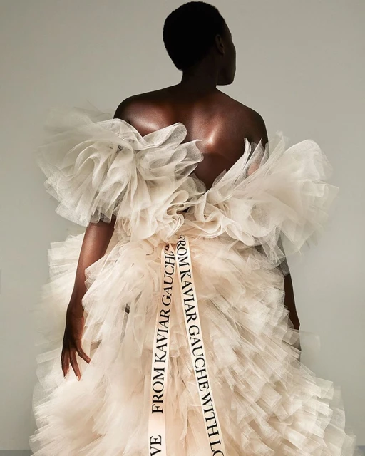 Back view of a woman in a voluminous ruffled dress with a ribbon belt that says 'From Kaviar Gauche with Love' against a plain background.